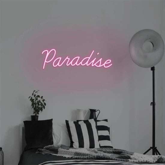 Pink Paradise neon sign mounted on wall above bed with striped pillows and floor lamp