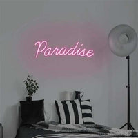 Pink Paradise neon sign mounted on wall above bed with striped pillows and floor lamp
