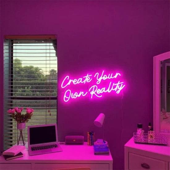 Create your own reality neon sign glowing pink on purple wall above desk with laptop and flowers